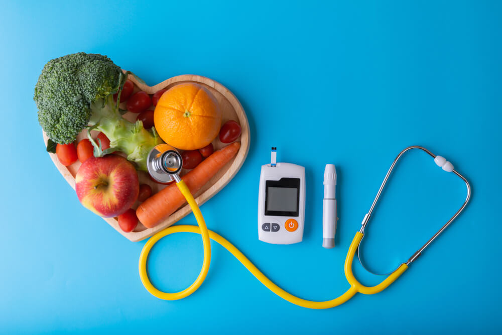 Fruits and vegetables in a heart shaped bowl and a stethoscope and diabetes monitor